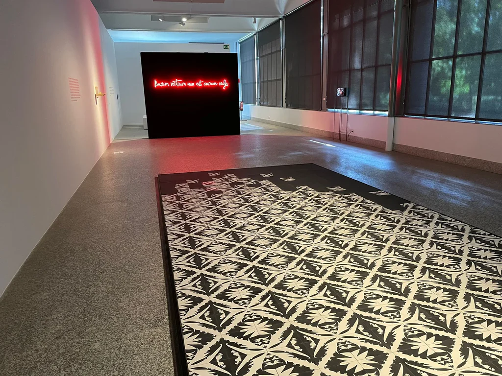 Alfombra de baldosa hidráulica en blanco y negro con dibujo geométrico repetido y orla oscura perimetral, colocada sobre un pavimento continuo en una sala de exposición